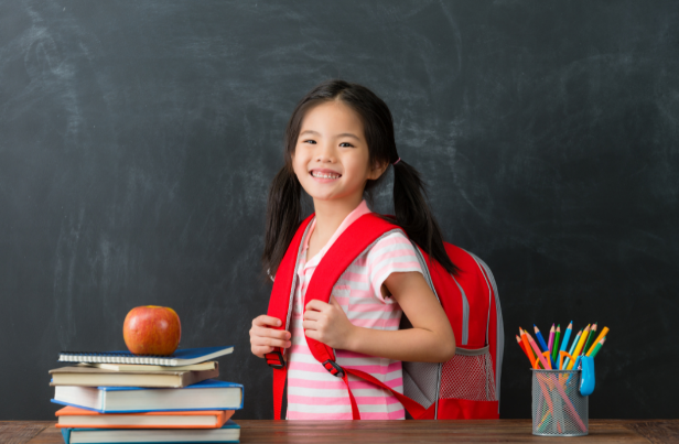 Young girl smiles behind a teachers' desk with her back pack on